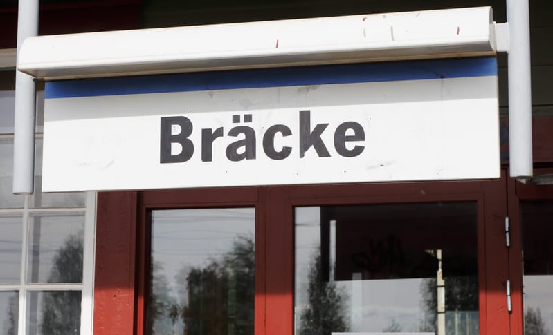 Bilden visar en vit skylt med texten "Bräcke" i svarta versala bokstäver. Ovanför texten finns ett blått streck. Skylten är fäst under ett vitt utskjutande tak eller en markis, som har en del smuts och enstaka röda streck. Under skylten syns en del a