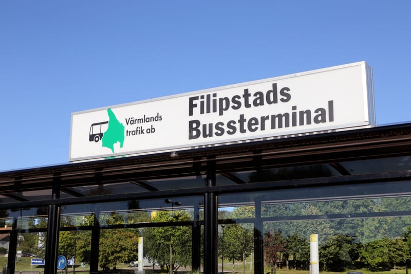 Bilden visar en skylt för 'Filipstads Bussterminal' under en klarblå himmel. Skylten är vit med svart text och inkluderar en logotyp med en stiliserad buss och en grön karta som representerar Värmlands trafik ab. Under skylten syns en del av en bussk
