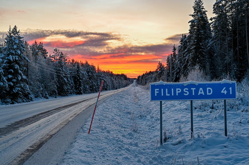 Bilden visar en vinterlandskap med en asfalterad väg som sträcker sig in i horisonten. Vägen är delvis täckt av snö och is. Vid sidan av vägen, till höger i förgrunden, står en blå vägskylt med texten "FILIPSTAD 41" i vit färg. Skylten har snö på ova