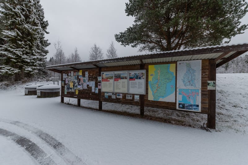 Bilden visar ett informativt anslagstavelsystem utomhus under vintertid. Den långa träkonstruktionen har ett tak med korrugerad plåt, som är täckt av ett tunt lager snö. På anslagstavlan finns diverse information, inklusive tidningsurklipp, kartor oc
