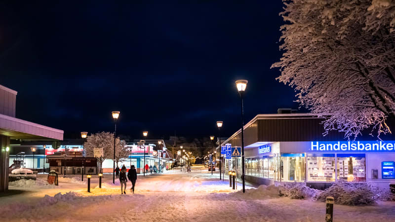 Bilden visar en snötäckt gatumiljö under en vinterkväll eller natt. Gatubelysningen är tänd och lyser upp den snöiga marken och de snöklädda träden. Till höger i bilden syns en byggnad med skylten "Handelsbanken" med blå belysning, samt en bankomat.
