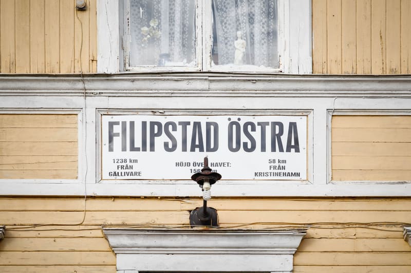 En detaljerad bild av en del av en gul träbyggnad med vita snickerier. I mitten av bilden finns en skylt med svart text på vit bakgrund där det står "FILIPSTAD ÖSTRA" överst. Under den texten finns mindre text som anger avstånd och höjd över havet. T