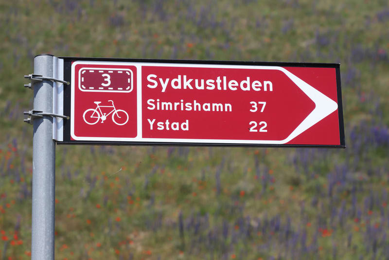 Bilden visar en röd vägskylt med vit text och symboler placerad på en grå stolpe. Skylten visar "Sydkustleden" med destinationerna "Simrishamn 37" och "Ystad 22". Till vänster på skylten finns en vit cykelsymbol och siffran "3" inramad i en streckad
