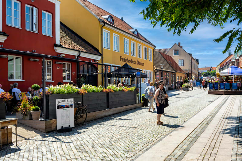 En gatuvy över en kullerstensgata kantad av flervåningsbyggnader i olika färger, inklusive rött och gult. På vänster sida finns en restaurang med en uteservering där flera personer sitter vid bord. Framför uteserveringen står en skylt med texten "Hic