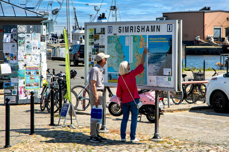 Bilden visar två personer, en man och en kvinna, som tittar på en stor informationstavla utomhus. Tavlan har en karta över omgivningarna och texten "SIMRISHAMN" överst. Kvinnan, klädd i en rosa blus och jeans, pekar på kartan. Mannen, med en keps och