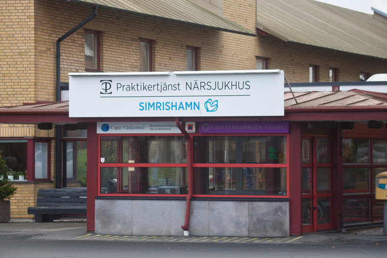 Bilden visar entréområdet till en byggnad, identifierad som "Praktikertjänst Närsjukhus Simrishamn" via en skylt ovanför ingången. Byggnadens fasad är av tegel i ljusare nyans. Entrépartiet har ett rött tak och röda fönsterramar. Under den stora skyl
