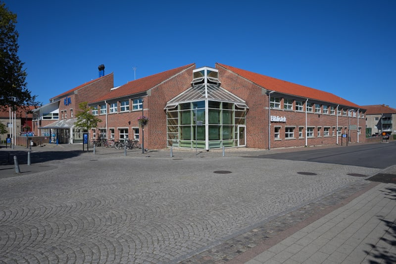 En exteriörbild tagen under dagtid, som visar en byggnad med fasader av tegel och tak av rött tegel. Byggnaden har flera fönster och en framträdande glaskonstruktion i mitten. Till vänster på byggnaden syns skyltar med texten "Hemköp" i blått. Till h