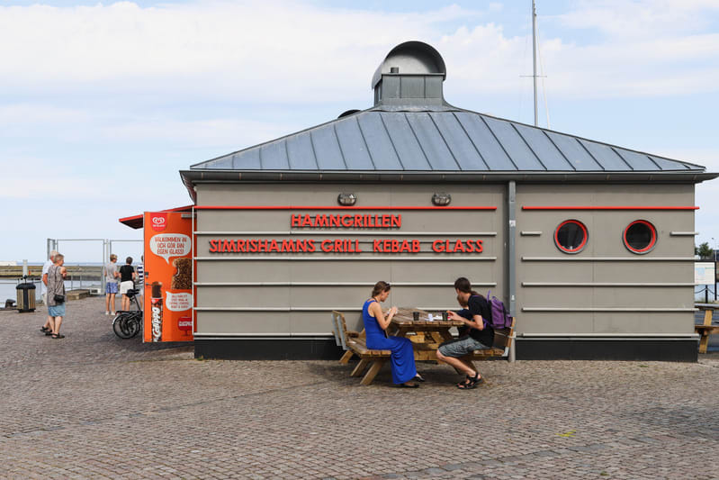 Bilden visar byggnaden "Hamngrillen Simrishamns Grill Kebab Glass" med text i rött på en grå fasad. Byggnaden har ett grått plåttak med en rundad kupa på toppen och två runda, röda fönster på sidoväggen. Framför byggnaden sitter två personer vid ett