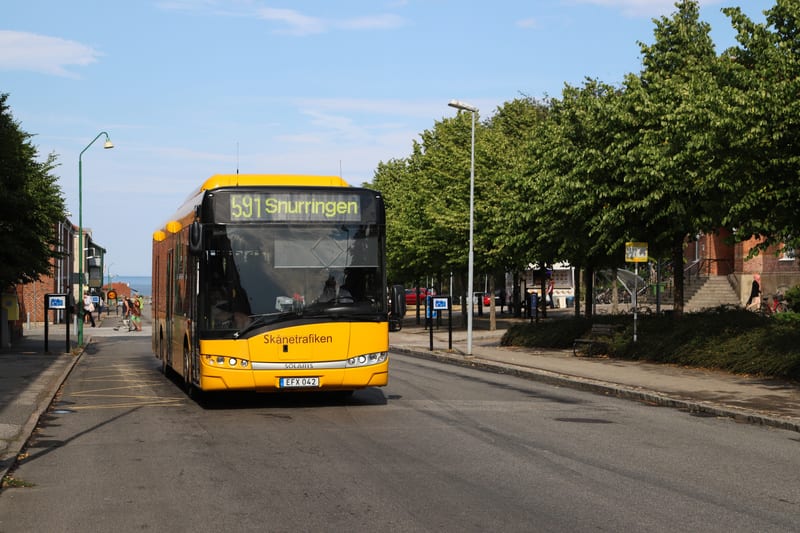 Bilden visar en gul buss från Skånetrafiken som står på en gata under dagtid. Bussen har destinationsskylten "591 Snurringen" synlig. I förgrunden syns en asfalterad väg med gula markeringar på asfalten till vänster om bussen. Till vänster om gatan f