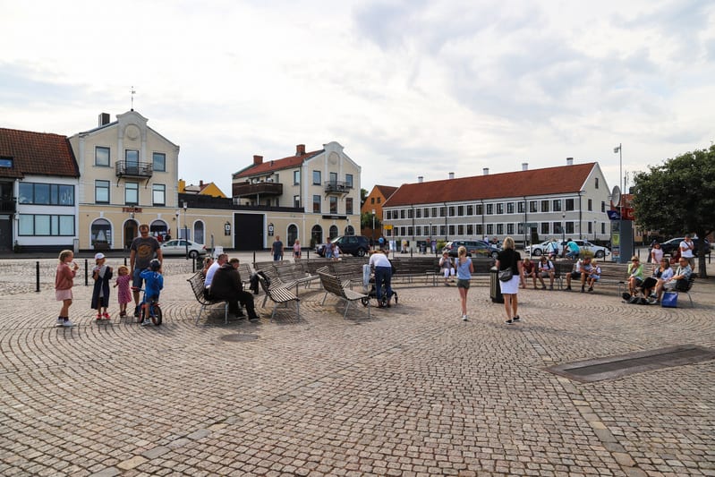 En utomhusbild som visar ett torg med kullersten. Flera människor syns i olika grupper; en familj med barn står till vänster, och andra sitter på bänkar utspridda över torget. I bakgrunden finns flera byggnader, varav några har röda tegeltak och ljus