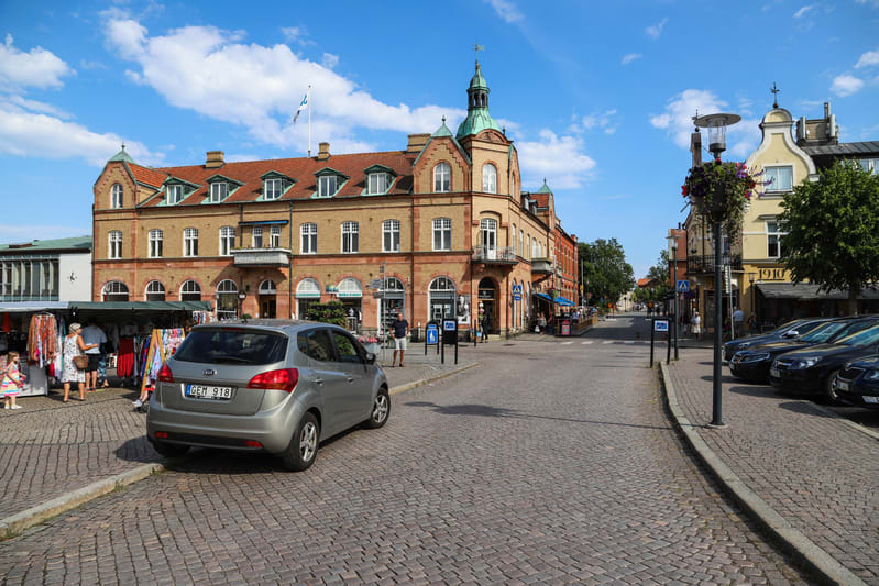 Bilden visar en gata i en stadsmiljö under dagtid. Gatstensbeläggning täcker gatan och torget. På vänster sida syns ett marknadsstånd med kläder och några personer som tittar på varorna. En silverfärgad bil är parkerad på gatstenen. I bakgrunden domi