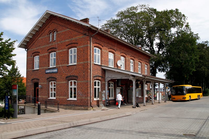 En bild tagen utomhus visar ett rött tegelhus, troligtvis en stationsbyggnad, med vita fönster. På byggnadens framsida finns skylten 'SIMRISHAMN'. En grå veranda med pelare sträcker sig längs ena sidan av huset mot en buss- eller tågperrong. En gul b