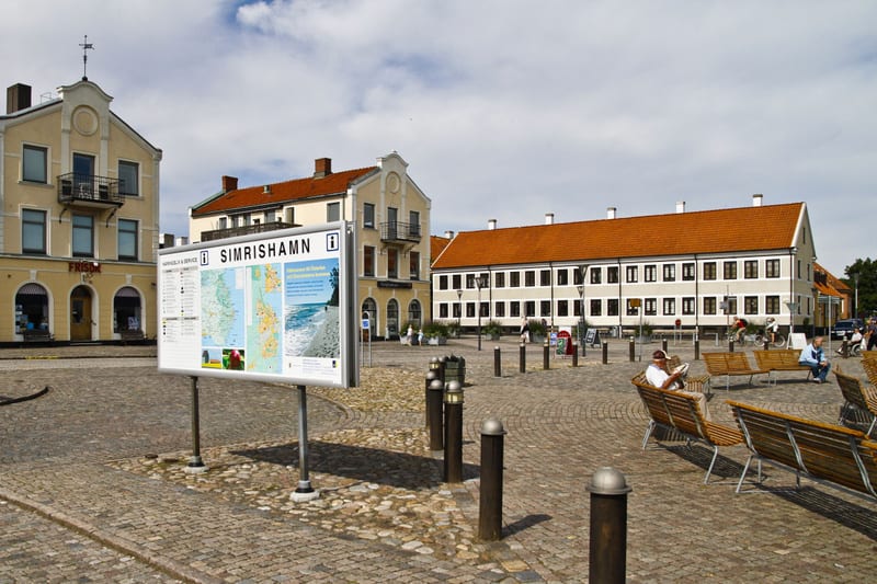 Bilden visar ett torg eller en öppen plats bebyggd med flera hus i bakgrunden. Ett skyltställ med texten "SIMRISHAMN" och kartor, samt annan information, står i förgrunden till vänster. Torget är belagt med gatsten och har flera träbänkar. Några pers