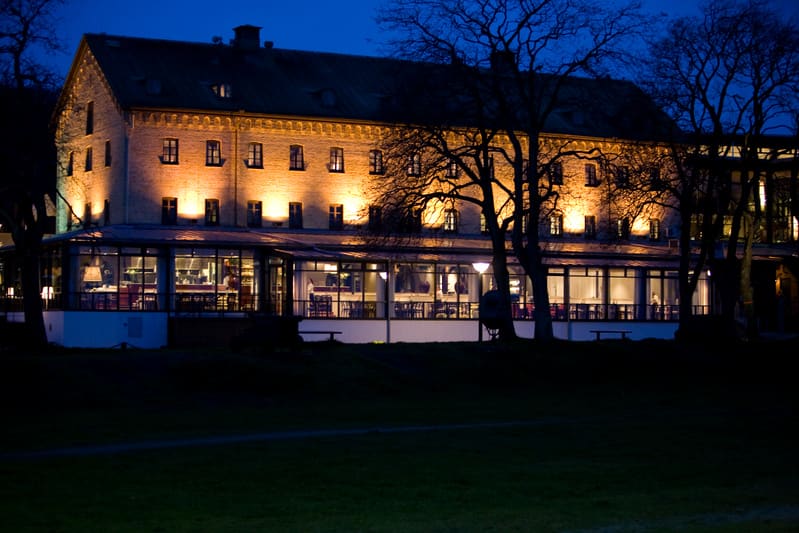 Bilden visar en stor byggnad i mörker, upplyst av flera spotlights som kastar gult ljus på fasaden. Byggnaden har flera våningar med fönster och en souterrängvåning med stora glaspartier som liknar en restaurang eller vinterträdgård. Genom glaspartie