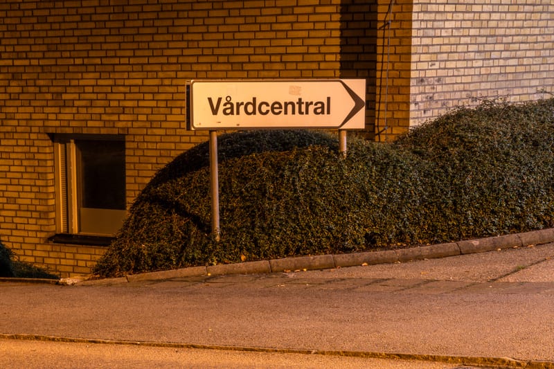 Bilden är tagen utomhus nattetid och visar en vit skylt med texten 'Vårdcentral' i svart, med en pil som pekar åt höger. Skylten är monterad på två metallstolpar och står framför en stor grön buske eller häck. Bakom busken och skylten syns en ljusbru