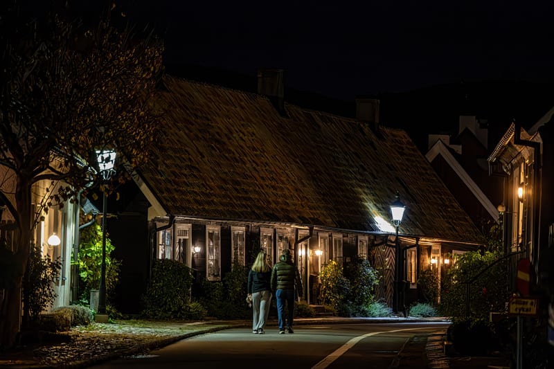 Bilden är tagen utomhus nattetid och visar en gata med flera hus i gammal stil. Två personer promenerar bort från kameran längs gatan, som har en vit heldragen linje. Husen är mörka med belysta fönster och dörrar, vilket indikerar att det är tänt ino