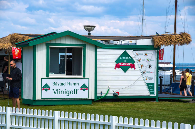 Bilden visar ett litet, vitt hus med grönt tak och gröna detaljer, märkt "Båstad Hamn Minigolf" på framsidan. Till höger om huset finns en anläggning för försäljning av churros och glass, vilket indikeras av skyltar med texten "CHURROS & GLASSCÁFET"