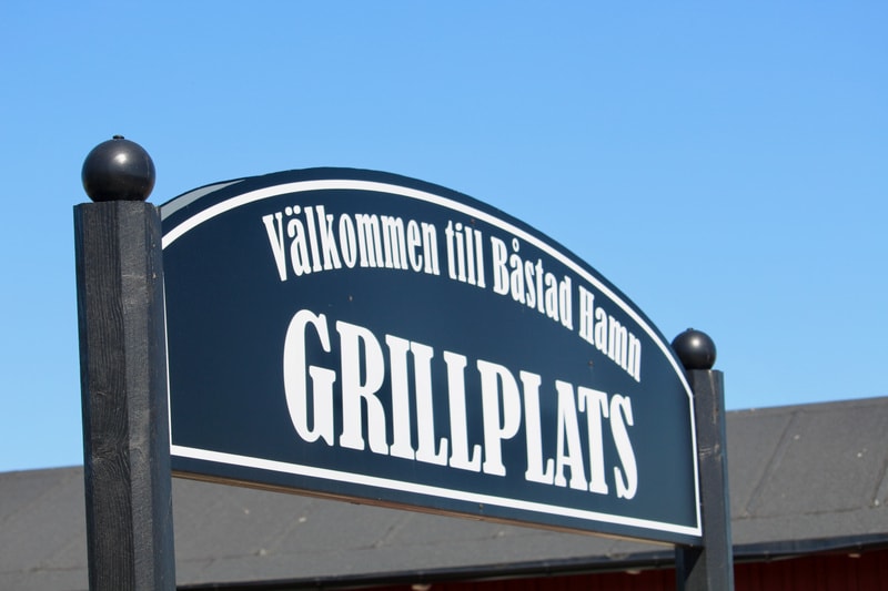 Bilden visar en skylt på nära håll med blå himmel i bakgrunden. Skylten är mörkblå med vit text och en vit ram. Texten längst upp på skylten lyder "Välkommen till Båstad Hamn" och under det står det "GRILLPLATS" i större bokstäver. Skylten är fäst vi