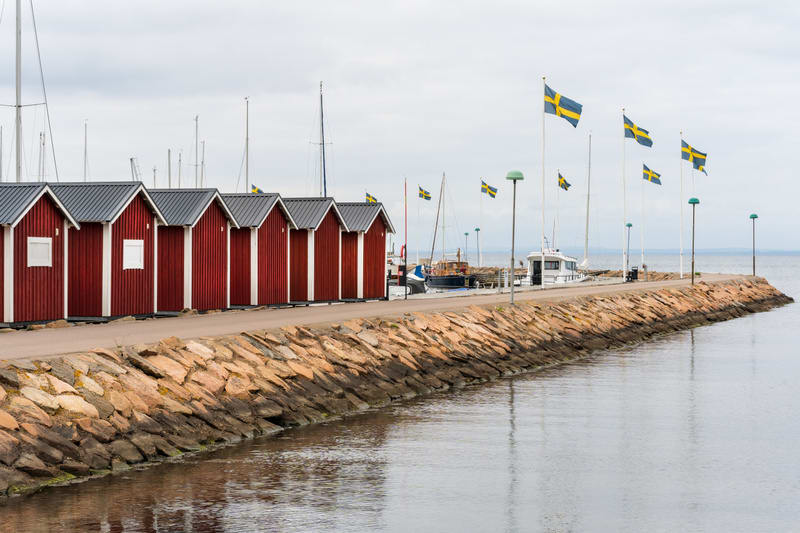En bild som visar en rad med röda sjöbodar placerade längs en kajkant. Mellan sjöbodarna och vattnet finns en stenmur. På kajen, bortom sjöbodarna, syns flera båtar förtöjda i en småbåtshamn. Ett flertal svenska flaggor vajar på höga flaggstänger län