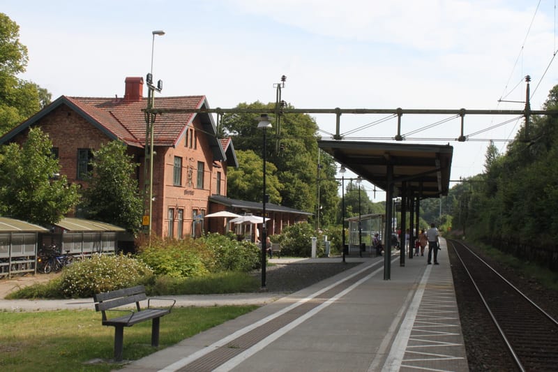 Bilden visar en järnvägsstation med ett tegelhus och en plattform på en solig dag. Till vänster i bild syns ett äldre tegelhus med rött tegeltak. Framför huset finns en uteservering med vita parasoller och stolar. Ett buskage med gröna löv och vita b
