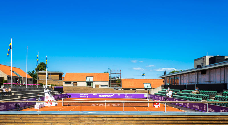 Bilden visar en utomhustennisbana i lera under en klarblå himmel. Runt tennisbanan finns gradänger med sittplatser. På långsidan av tennisbanan syns ett staket med reklam för olika företag, samt texten "ATP WORLD TOUR". I bakgrunden syns byggnader me