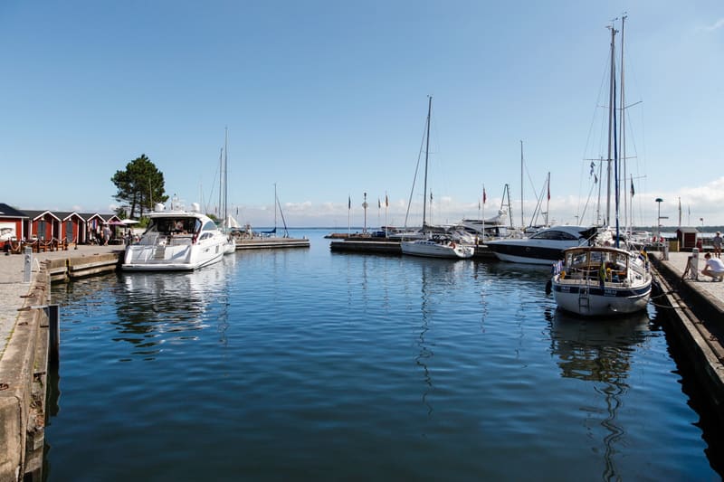 Bilden visar en småbåtshamn med flera förtöjda båtar i klart, blått vatten. I förgrunden till vänster syns en stor vit motorbåt och till höger en segelbåt med namnet "ARAR" synlig på aktern. Längs vänster sida av hamnen syns röd-vita sjöbodar eller s