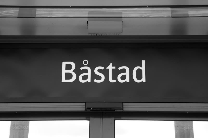 Bilden visar en närbild på en mörk skylt med vit text där ordet "Båstad" står skrivet i versaler med inledande stor bokstav. Skylten är placerad ovanför vad som verkar vara en dörröppning, där överkanten av två dörrblad syns tydligt i nederkant av bi