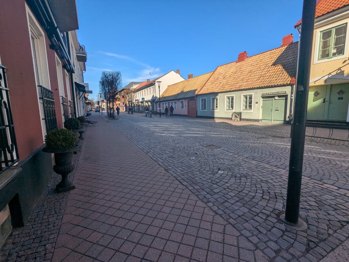 Bilden visar en gata i en stadsmiljö under dagtid. Gatstensbeläggning täcker vägbanan och en del av trottoaren, medan den andra delen av trottoaren består av fyrkantiga stenplattor. Längs gatan står byggnader i olika färger som rött, gult, ljusgrönt och rosa, med tak av tegelpannor i orangea nyanser. Flera hus har grönmålade fönsterkarmar och dörrar. På vänster sida syns husfasader med fönster dekorerade med räcken och krukor med gröna växter framför. I fjärran kan man se träd med kala grenar och en klarblå himmel ovanför. Några personer promenerar längre bort på gatan och en cyklist syns i mitten av bilden. Solen kastar långa skuggor över marken.
