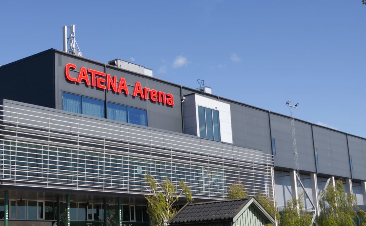 Bilden visar byggnaden "CATENA Arena" under en klarblå himmel med några vita moln. Byggnaden har en modern design med grå fasad och stora fönsterpartier. Ovanför de nedre fönstren finns en horisontell struktur med vita lameller. Ett stort, rött lysan