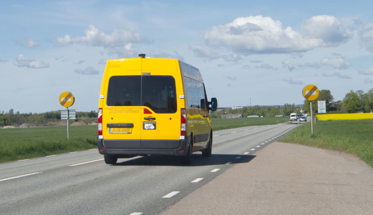 Bilden visar ett gult fordon, en minibuss, som kör på en landsväg. Fordonet är märkt med texten "Skånetrafiken" på bakdelen. Vägen har vita körfältsmarkeringar. Längs vägen syns gräsbevuxna områden och träd i bakgrunden. På höger sida av vägen, längr