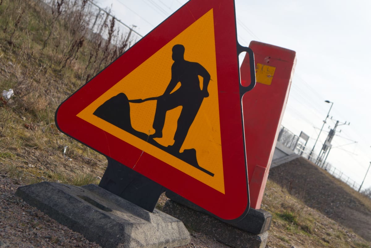 En gul varningsskylt med röd kant, föreställande en svart silhuett av en person som skottar, står lutad i bilden. Skylten är fäst vid en röd avlång stolpe och är placerad nära marken, som består av grus och torrt gräs. I bakgrunden syns en grönskande