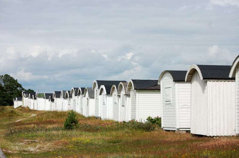En bild tagen utomhus som visar en lång rad av vita strandhytter med svarta, välvda tak. Hytterna sträcker sig diagonalt från vänster till höger i bilden och minskar i storlek ju längre bort de är. Framför hytterna finns ett område med gräs och vissn