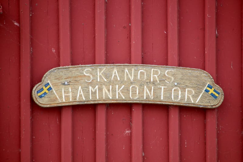 Bilden visar en träskylt med texten "SKANÖRS HAMNKONTOR" inristad. Skylten har en böjd form och är fäst vid en röd träpanelvägg med vertikala brädor. På vardera sida om den inristade texten finns avbildade svenska flaggor. Texten är huggen i ljusare