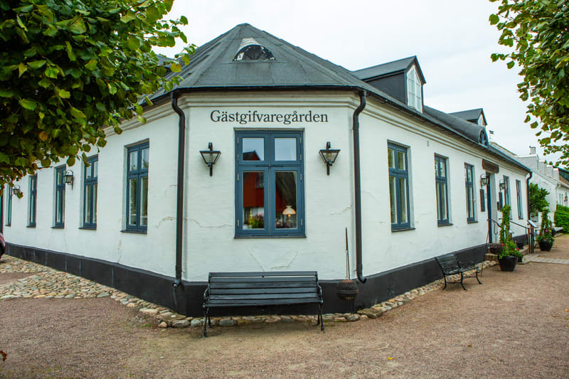 Bilden visar fasaden på en vit byggnad med svarta fönsterkarmar och en mörk takfot. På fasaden står det 'Gästgifvaregården' skrivet. Fönster är synliga, varav ett stort fönster i mitten av bilden har en synlig lampa inuti. Framför byggnaden finns en