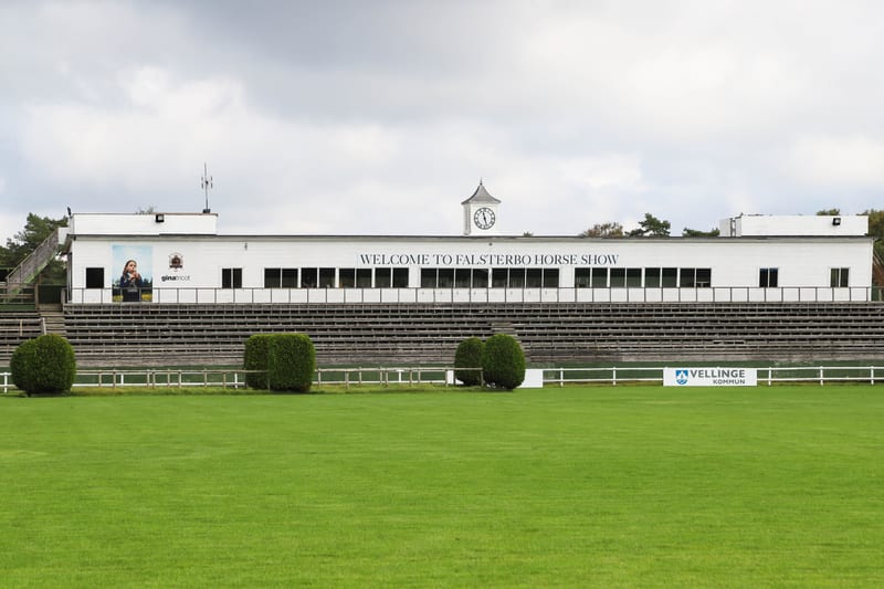 Bilden visar Falsterbo Horse Show-läktaren och det omgivande gräsplanen under en molnig dag. Läktaren är en vit byggnad med ett klocktorn i mitten och texten 'WELCOME TO FALSTERBO HORSE SHOW' stort skrivet på fasaden. Under den vita byggnaden finns f
