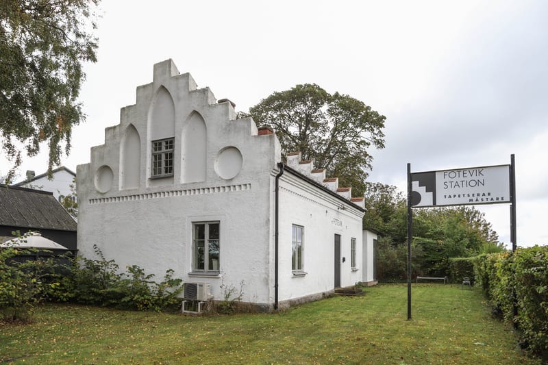 En vit, låg byggnad med flera fönster under en mestadels gråmolnig himmel. Byggnaden har en iögonfallande trappgavel med flera utskjutande steg och dekorativa, valvbågsformade fördjupningar samt cirkulära detaljer. Väggarna är putsade och fönsterkarm