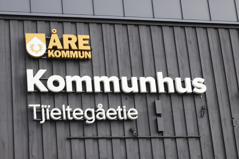 Bilden visar en del av en byggnads exteriör med skyltar. Väggen är klädd med mörkgrå träpanel. Överst till vänster syns en skylt med guldgul text som lyder "ÅRE KOMMUN" och bredvid texten finns kommunvapnet i gult och vitt. Nedanför kommunens skylt f
