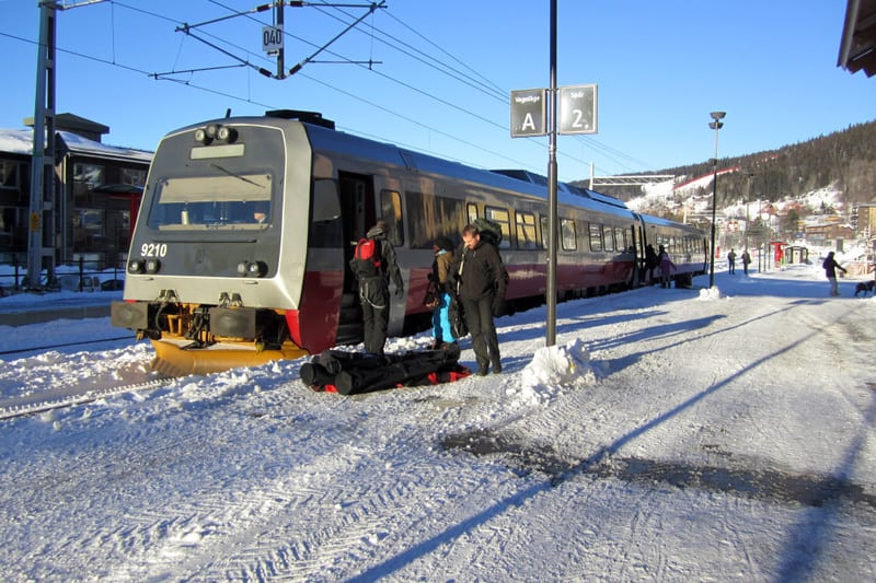 En bild som visar ett tåg som står vid en perrong på en snötäckt tågstation under dagtid på vintern. Flera personer står på perrongen, dels nära tågets öppna dörr där några verkar kliva på eller av, och dels längre bort på perrongen. Tåget är grått m