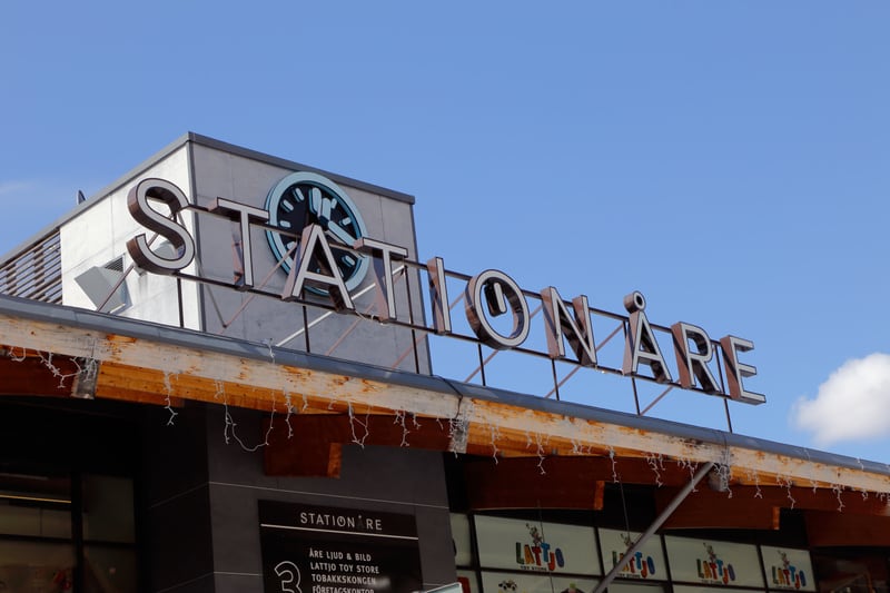 Bilden visar delar av fasaden på en byggnad under en klarblå himmel med några vita moln. I förgrunden syns en stor, belyst skylt med texten "STATION ÅRE". Bokstäverna är i metall med en tredimensionell effekt och de har en ljuskälla bakom sig som lyse