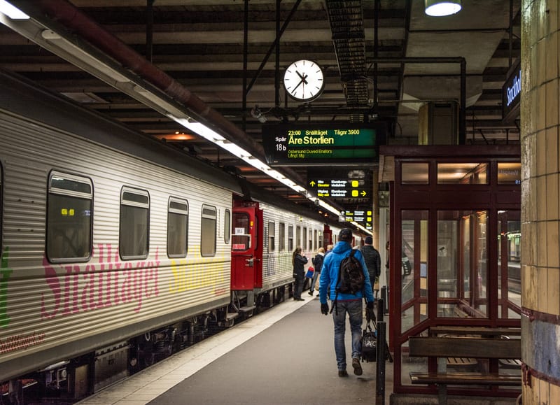 Bilden visar en järnvägsperrong inomhus med ett tåg som står vid spåret. Människor syns på perrongen, bland annat en person i blå jacka och ryggsäck som går bort från kameran. Tåget är silverfärgat med röda detaljer och har synlig graffiti i rosa, gu