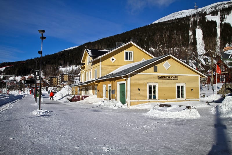 Bilden visar byggnader i ett snötäckt landskap under dagtid med klarblå himmel. Huvudfokus är en ljusgul byggnad med texten "BAHNHOF CAFÉ" på fasaden. Byggnaden har flera fönster och en grön dörr till vänster om entrén. Framför byggnaden finns ett sn