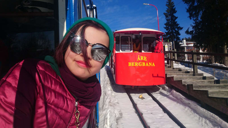 En person tar en selfie från insidan av vad som verkar vara en lift eller vagn på en bergbana. Personen bär en lila dunjacka, en vinröd halsduk och en turkos mössa, samt solglasögon som reflekterar omgivningen. Bredvid personen, utanför vagnen, syns