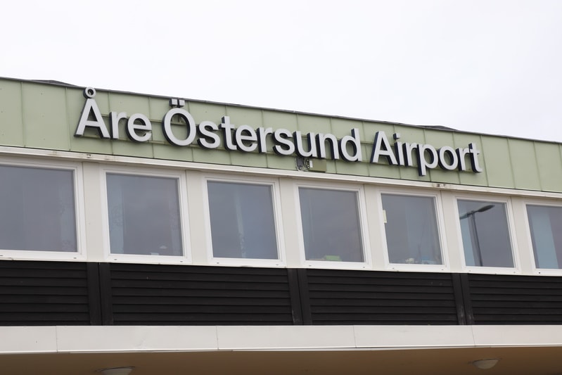 Bilden visar fasaden på en byggnadsdel med gröna paneldetaljer upptill och svarta paneler nedtill. Ovanför de svarta panelerna finns en rad vita fönsterkarmar med reflekterande glas. Ovanför fönstren, på den gröna panelen, sitter stora vita bokstäver