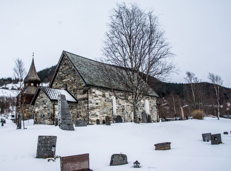En bild visar en stenskyrka med ett spetsigt torn, omgiven av snö. Kyrkan har ett grått tak och väggar av oregelbundna stenar. Flera gravstenar sticker upp ur snön runt kyrkobyggnaden. I bildens centrum står ett kalt träd intill kyrkan. I bakgrunden
