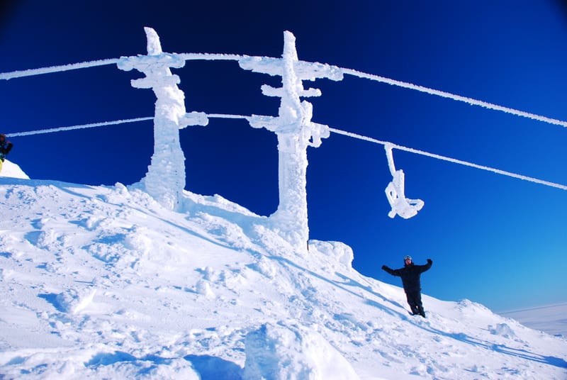 Bilden visar ett snötäckt, bergigt landskap under en klarblå himmel. Mitt i bilden syns en skidlift med två stänger täckta av snö och is. Mellan stolparna sträcker sig vajrar som också är täckta av is, och en ensam skidliftstol är täckt av frost och