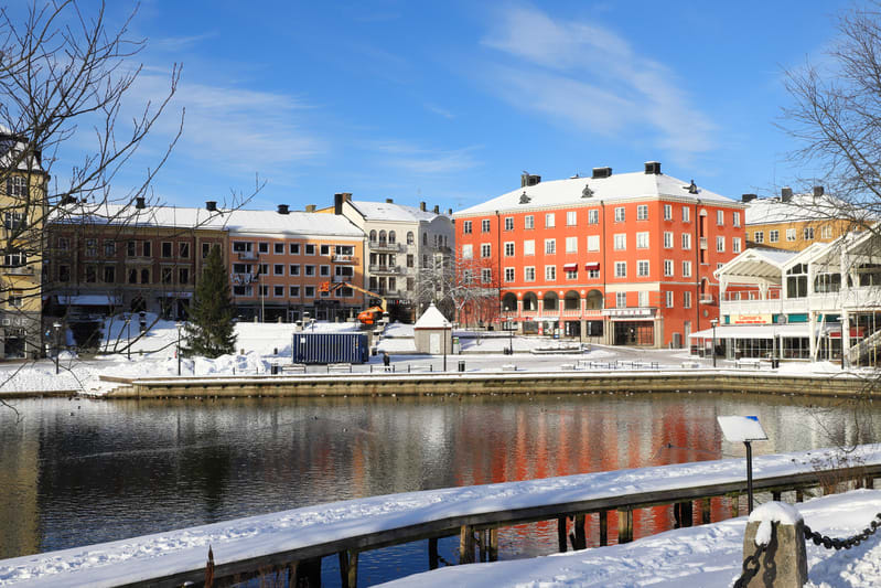 Bilden visar ett vinterlandskap med byggnader, vatten och snö. I förgrunden syns vatten som reflekterar de färgglada byggnaderna i bakgrunden. Kusten är täckt av snö, och en träbrygga med snö på ligger vid vattnet. På land syns flera byggnader i olik