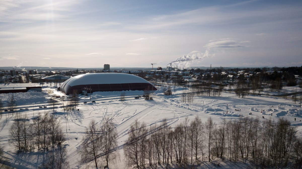 Bilden visar ett snötäckt landskap med en stor, kupolformad byggnad i förgrunden, delvis täckt av snö. Byggnaden har en rödbrun bas och en vit ovansida. I bakgrunden syns en stad eller tätort med flera byggnader, inklusive högre hus och industrianläg