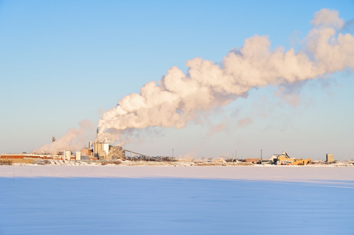 Bilden visar ett industriområde på avstånd en solig vinterdag. I förgrunden syns ett stort, snötäckt fält som sträcker sig över hela bildens bredd. Marken är täckt av en jämn snöyta. I bakgrunden reser sig ett större industrikomplex med flera byggnad