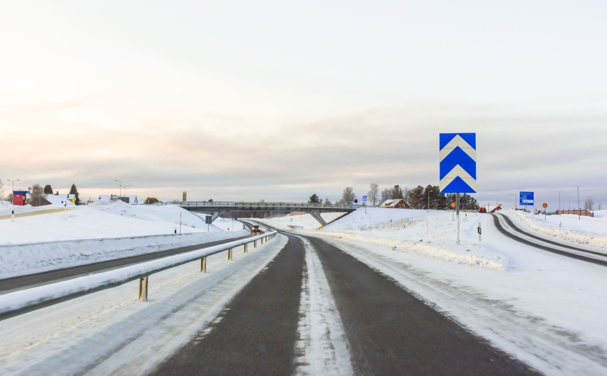 En bild tagen från ett fordon som visar en vinterlandskapsvy av en väg i norra Sverige. Vägen är mestadels täckt av snö på sidorna, med asfalterade körfält som har vita linjer. En stor blå och vit vägskylt med chevronsymboler (omväxlande blå och vita