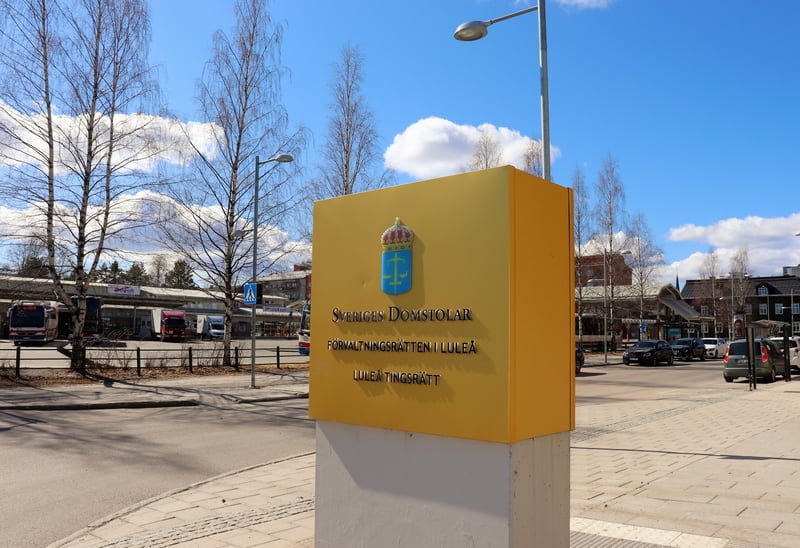 Bilden visar en gul skylt med texten "Sveriges Domstolar Förvaltningsrätten i Luleå Luleå Tingsrätt" och Sveriges Domstolars emblem. Skylten står på en trottoar med grå stenplattor. I bakgrunden till vänster syns flera träd utan löv, busshållplatser